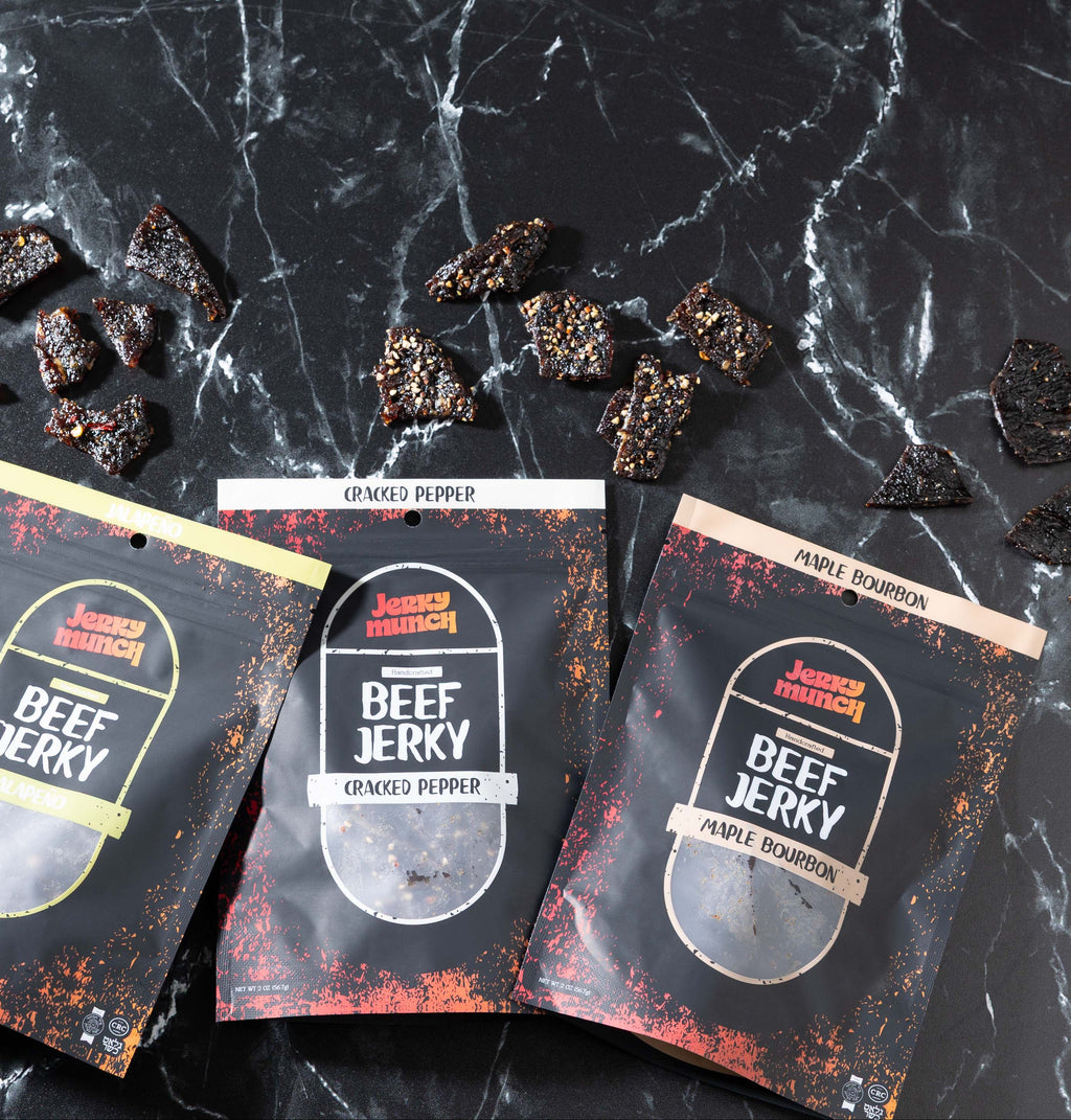 Three packages of Jerky Munch Kosher beef jerky on a black marble surface with scattered pieces of jerky.